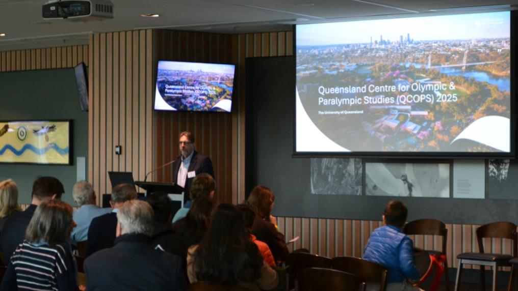 A speaker addresses an audience at a conference. A large screen behind the speaker shows an aerial view a city, while attendees watch from their seats.