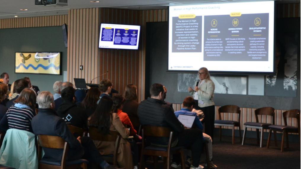 A presenter stands at the front of a conference room speaking to an audience. A large presentation screen is behind her, while attendees sit facing the stage.