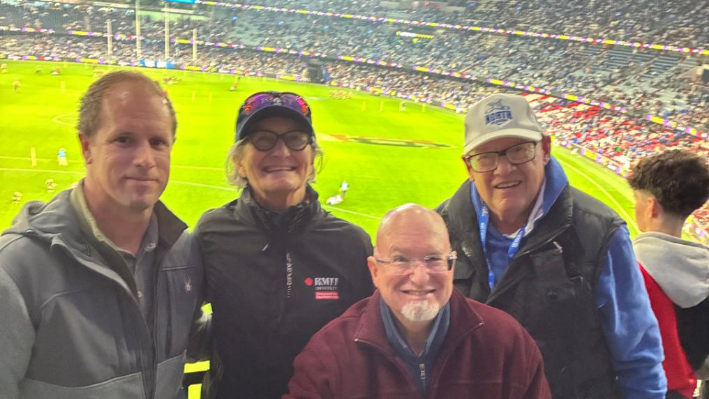 Four attendees posing in a stadium seating area overlooking a brightly lit AFL sports field. Rows of spectators fill the background.