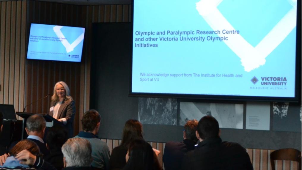 A presenter stands at the front of a conference room speaking to an audience. A large presentation screen displays a Victoria University slide, while attendees sit facing the stage.