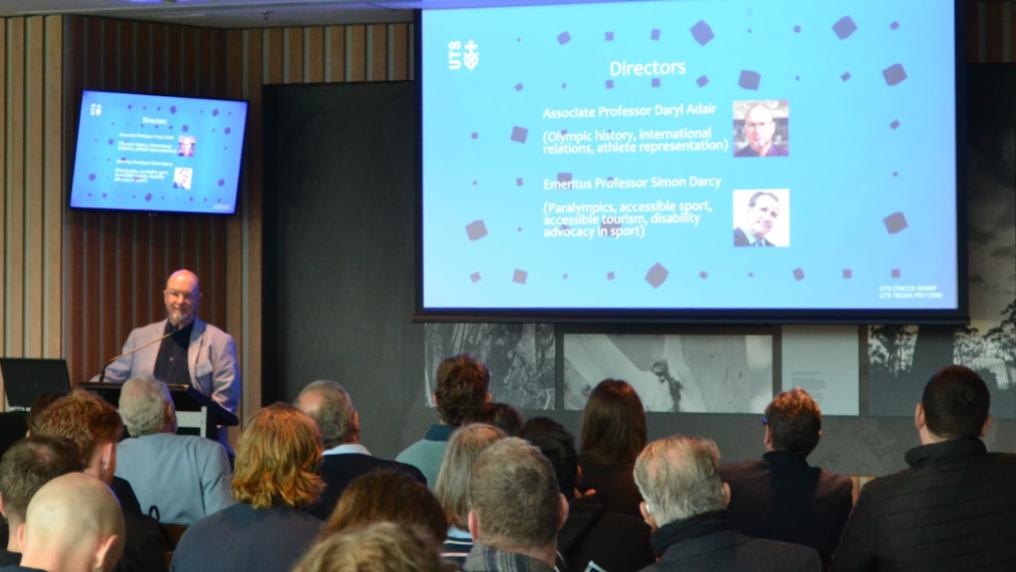 A speaker addresses an audience at a conference. A large screen behind the speaker shows a Victoria University presentation slide, while attendees watch from their seats.