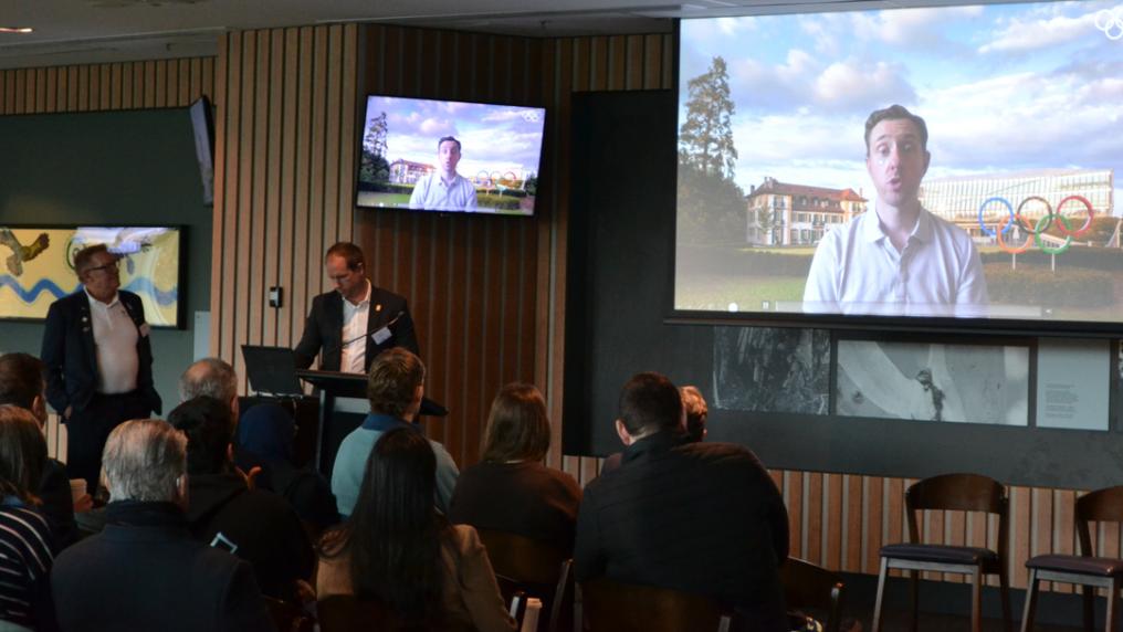 A large screen at a conference venue shows a person in business attire speaking, while attendees watch from their seats.