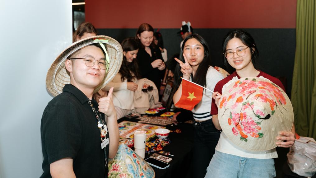 People gathered around a table at an indoor cultural and social event. Participants display decorated conical bamboo hats and a small flag while others socialise in background.