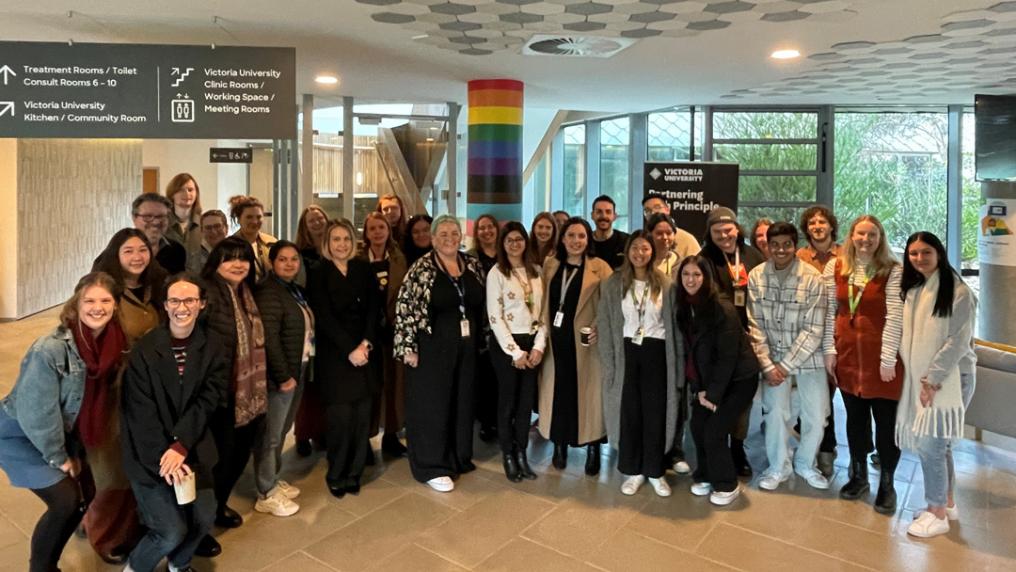 A large happy group of young adults in a foyer with a rainbow pride symbol and Victoria University signage