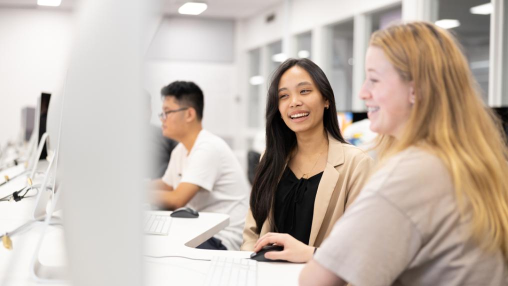 Students in a bright computer lab with large, modern, white computer monitors