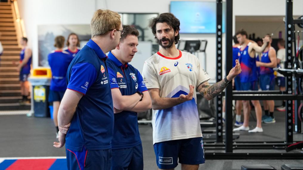 Three people are standing inside a busy gym. One is explaining something and the other two are listening. In the blurred background there are other figures of people in sports clothing and gym equipment.
