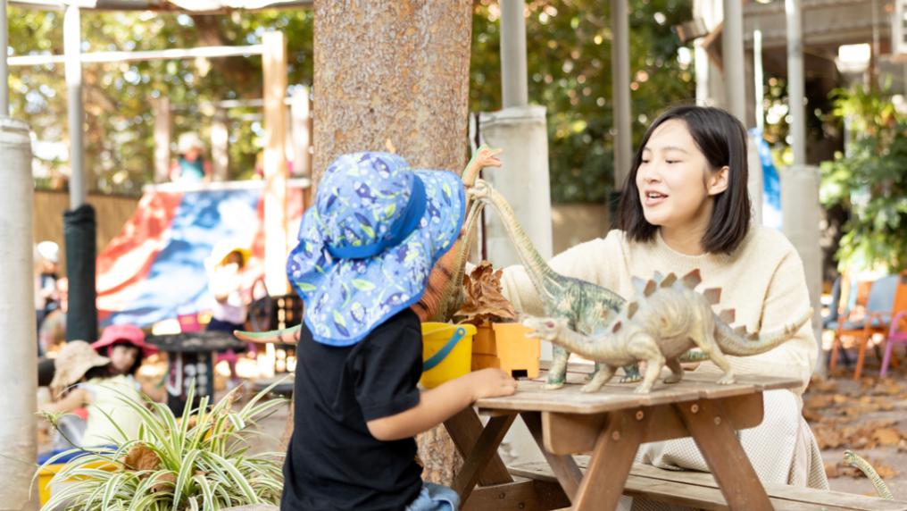 A young teacher interacts with a child in a bright outdoor space with toys, plants and play equipment