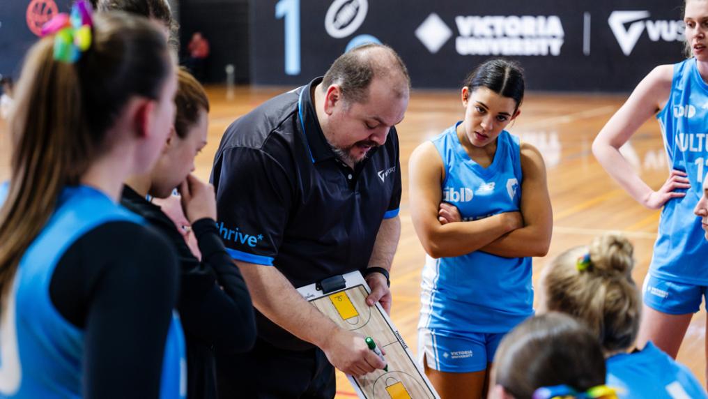 A woman's basketball team in VU blue uniforms huddles around a coach with clipboard