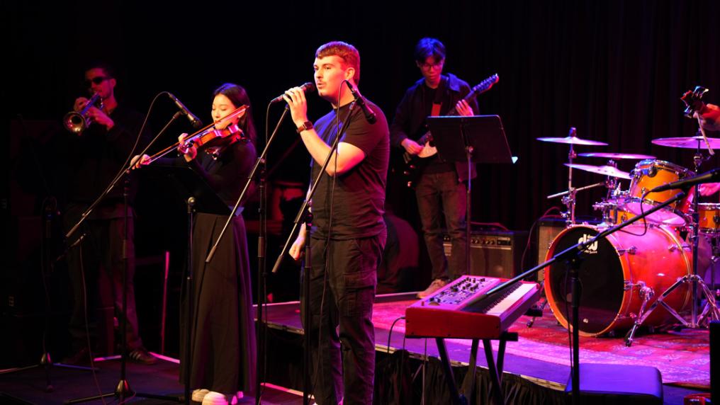 A young musical group with drums, vocal, violin & trumpet perform on a darkened stage with violet lighting