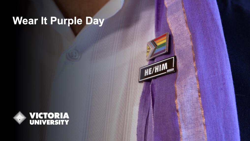 Close up of a person's white shirt and purple scarf, with LGBTQI flag and he/him badges. Text says 'Wear it purple day' and Victoria University logo. 