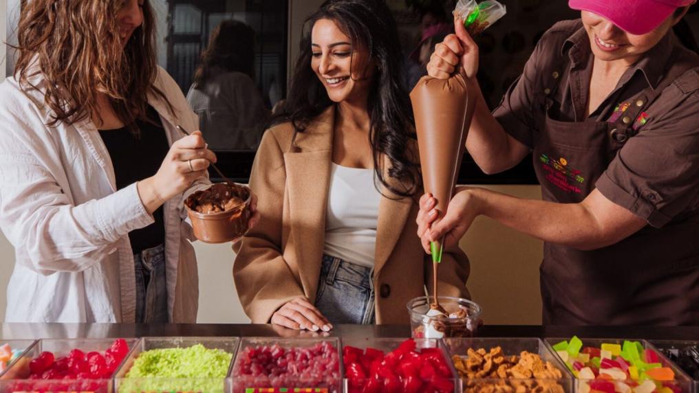 2 young women having fun at a lolly bar, while a worker in uniform and apron pours chocolate