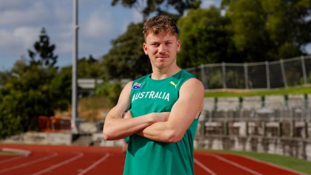 A young male athlete in Aussie green poses arms crossed on a running track
