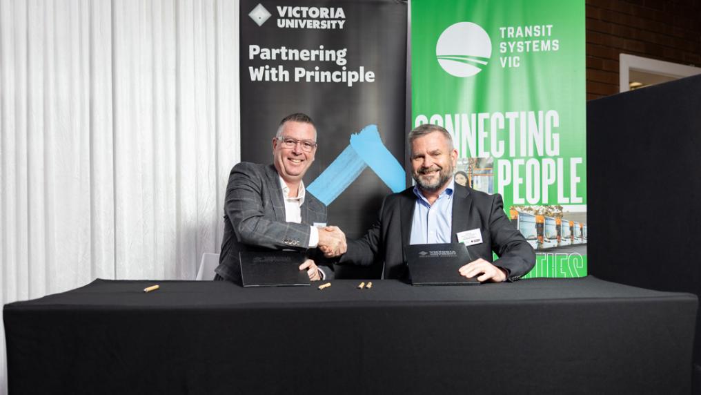 Two men in suits shake hands in front of Victoria University and Transit banners