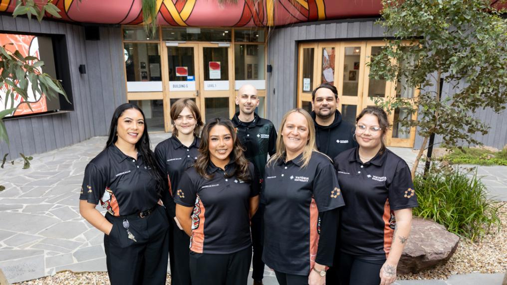Group of 7 smiling people posing outside with university building and trees in background. They are wearing matching black polo shirts with small Victoria University logo and some bright aboriginal artwork designs.