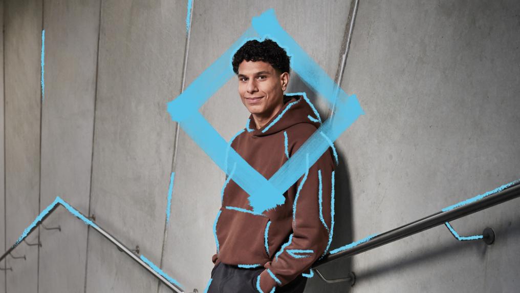A young man in casual brown hoodie is leaning against a concrete wall of a staircase. He is smiling and relaxed with his hands in his pockets. There is a blue diamond graphic around his head and upper body.