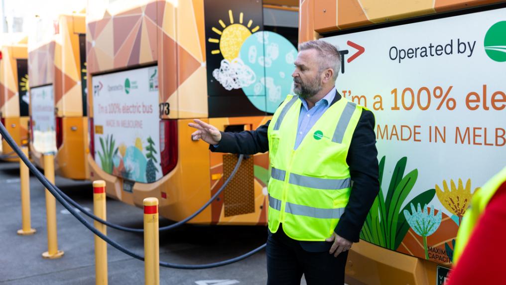 A middle-aged man in a suit and hi-vis jacket in front of a row of orange buses