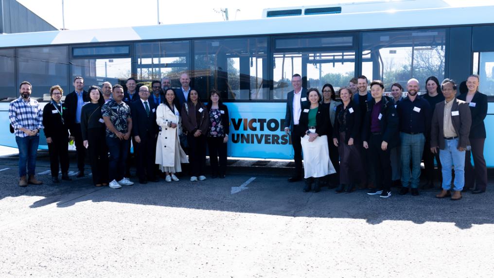 A large group of professional looking adults stands in front of a Victoria University branded bus