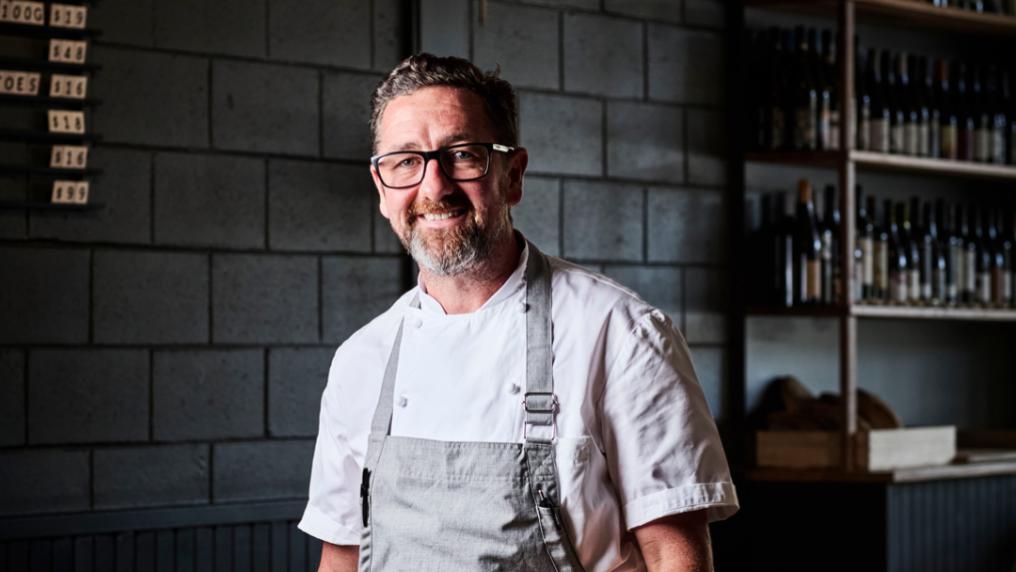 A chef in a clean white uniform in apron in a sophisticated food prep area with shelves of wine