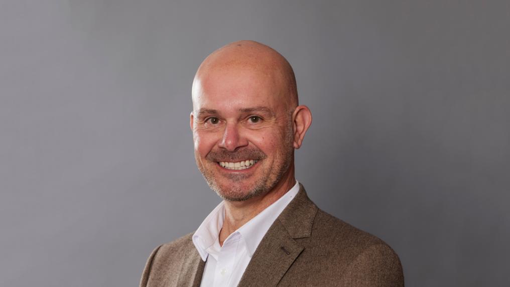 Professional looking man in brown jacket and white shirt smiles at the camera.