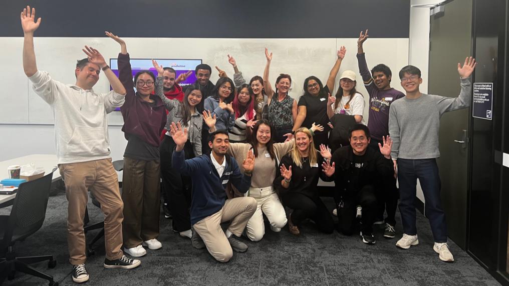 Large group of smiling people in casual dress wave at camera. They are inside a large meeting room.