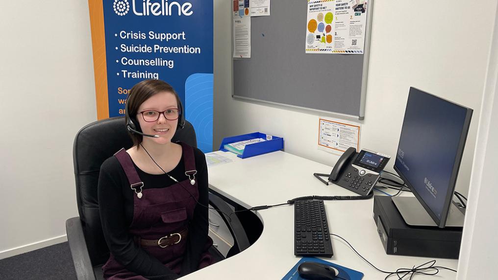 Young woman in casual clothing, wears a microphone headset at an office desk with Lifelife poster