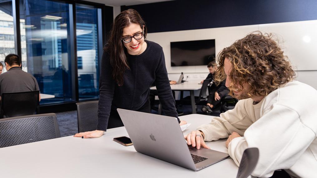 Young man at a desk in a modern classroom shows a smiling woman in glasses something on a laptop