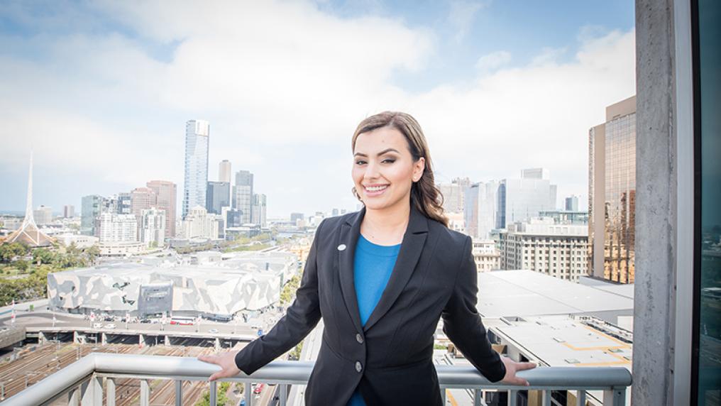 A young woman in professional dress stands on a city balcony with a cityscape behind her