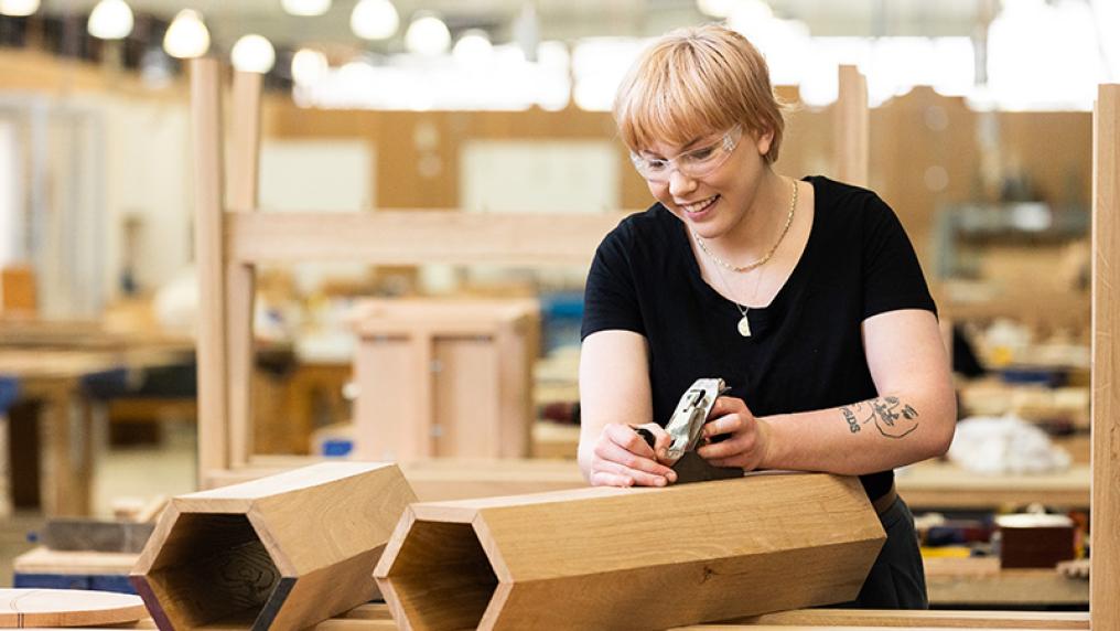  A young woman smiles as she planes a hexagonal woodwork creation in a large workshop