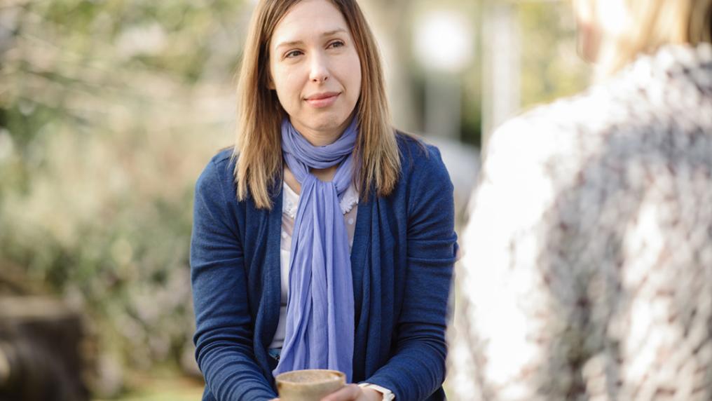 A middle aged woman in a purple scarf looks serious as she speaks with someone