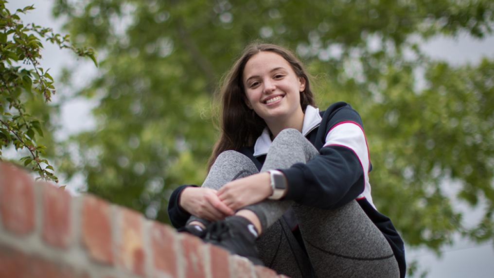 Elise Mercieca sitting smiling on top of a brick fence in front of a large tree