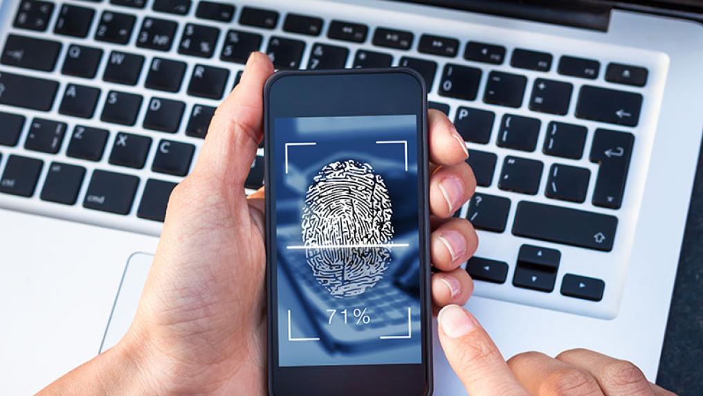  Hand holding a mobile phone in front of a laptop, with a security fingerprint icon on it.