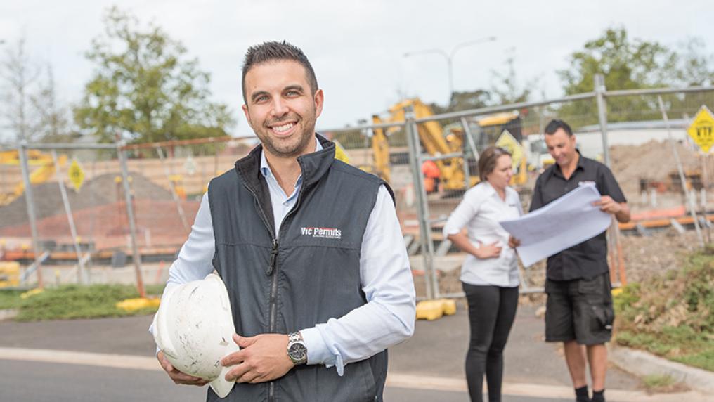 Michael Tabone in a Vic Permits vest, in front of a construction site, holding a hard hat, with two people behind him looking at paperwork