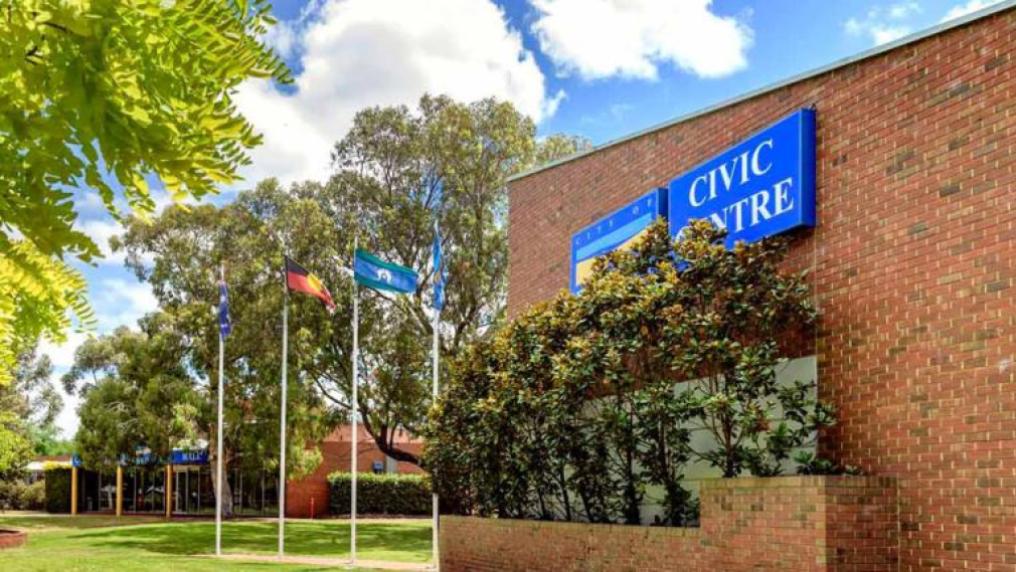 A brick building with a blue sign that reads 'Civic Centre', on a lawn with Australian flags in the background