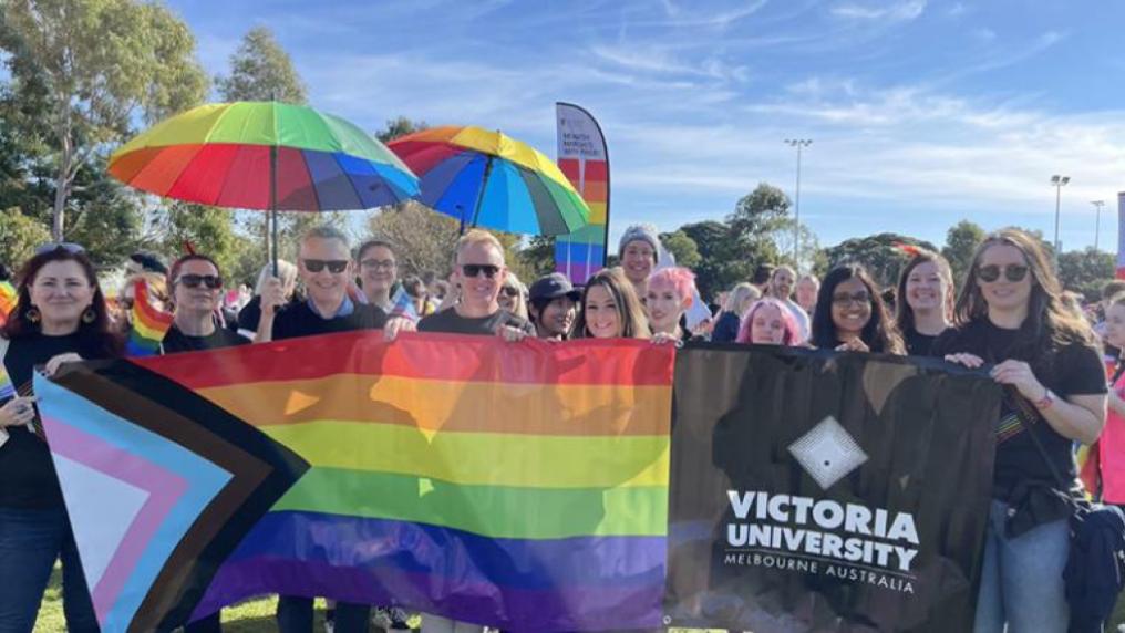 Group of men and women holding rainbow umbrellas, flag with Victoria University logo, at an outdoor gathering