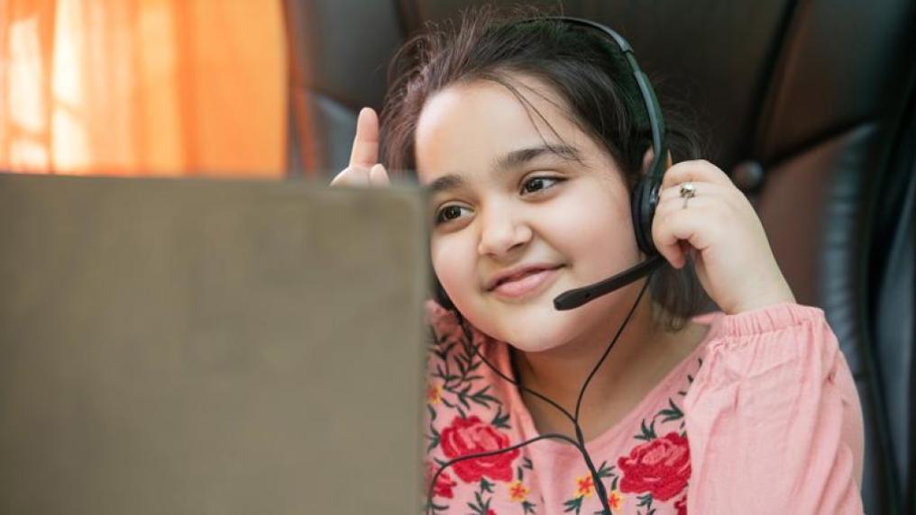 a school-aged girl with a headset sits in front of a computer with a hand raised