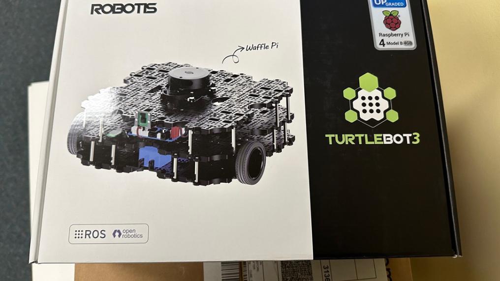A closed cardboard box. The box has a picture of a small square robot with wheels. The box says "Robotis" and "Turtlebot3" with a Raspberry Pi logo.