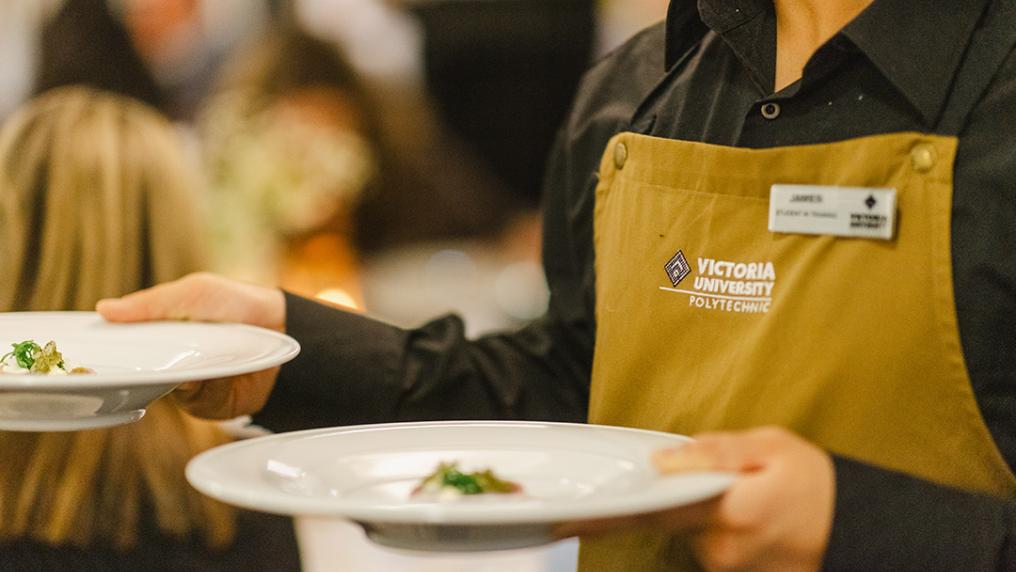 Wait staff in a Victoria University apron carries sophisticated looking dish