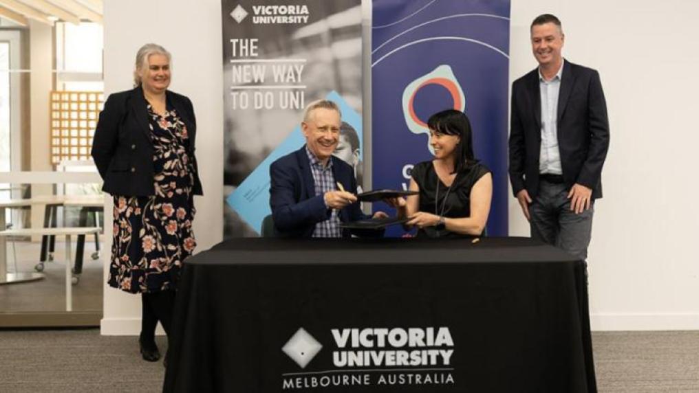 A man and woman smile at a Victoria University branded table, with branded banners in the background and two smiling, professional looking people