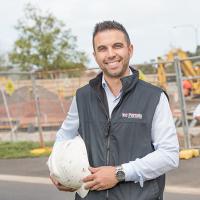 Michael Tabone in a Vic Permits vest, in front of a construction site, holding a hard hat, with two people behind him looking at paperwork