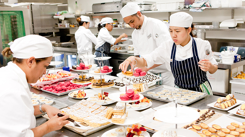 Chefs in a large industrial kitchen preparing an assortment of pastries.